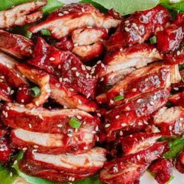 Sliced Chinese BBQ pork with red glaze, sesame seeds, and green onions on lettuce leaves.