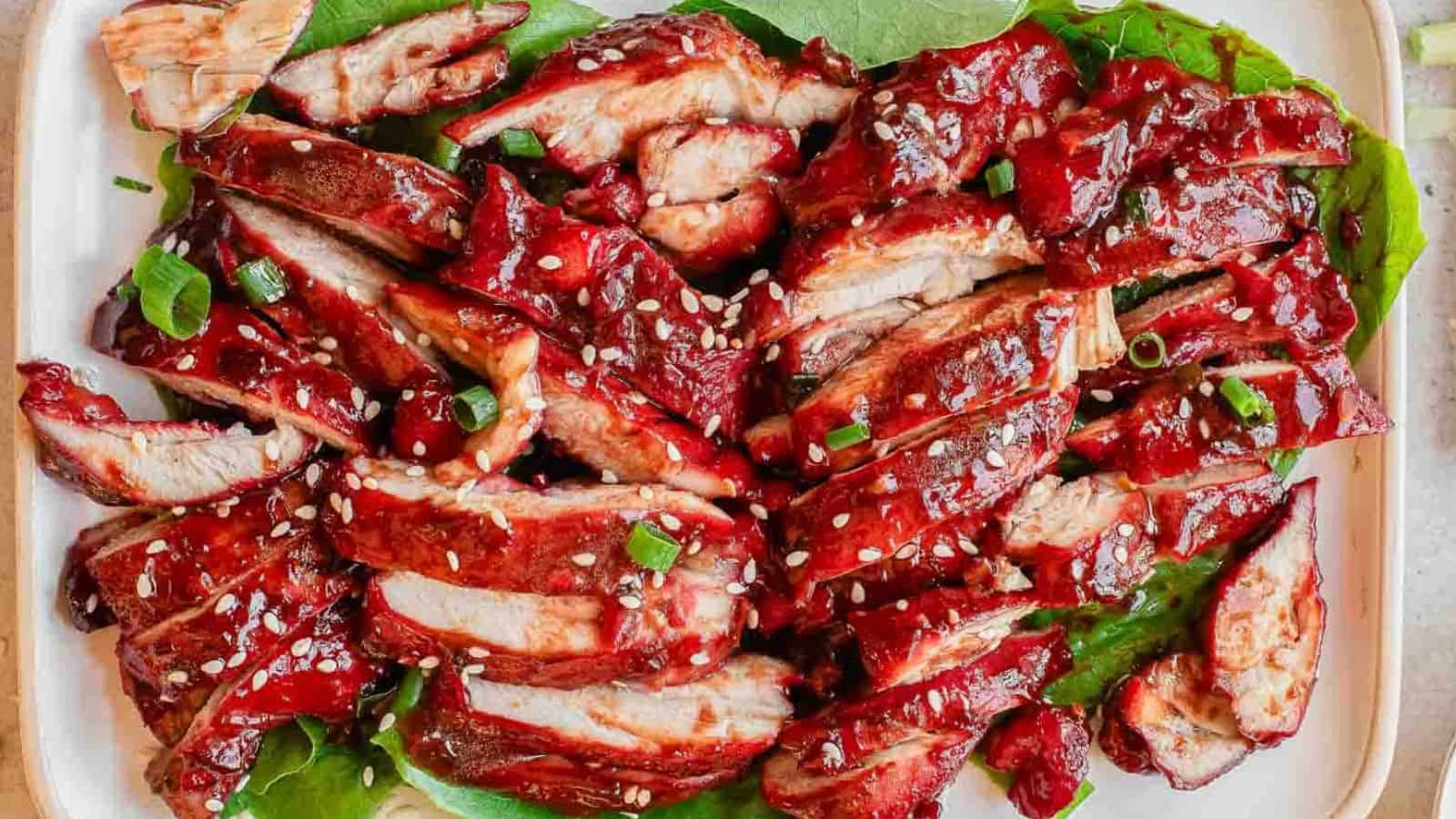 Sliced Chinese BBQ pork with red glaze, sesame seeds, and green onions on lettuce leaves.