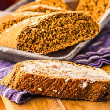 A slice of brown bread with butter sits in front of a tray of sliced bread on a purple cloth.