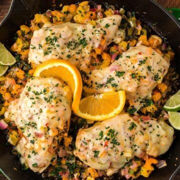 Chicken breasts in a skillet with melted cheese, diced veggies, and garnished with orange and lime slices.