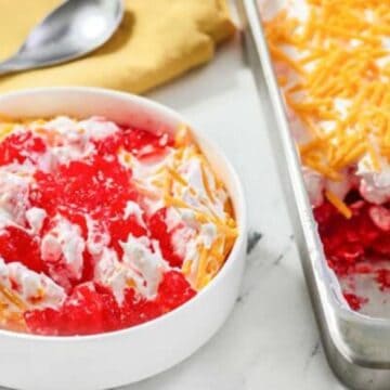 A bowl and pan of red gelatin salad topped with whipped cream and shredded cheddar cheese.
