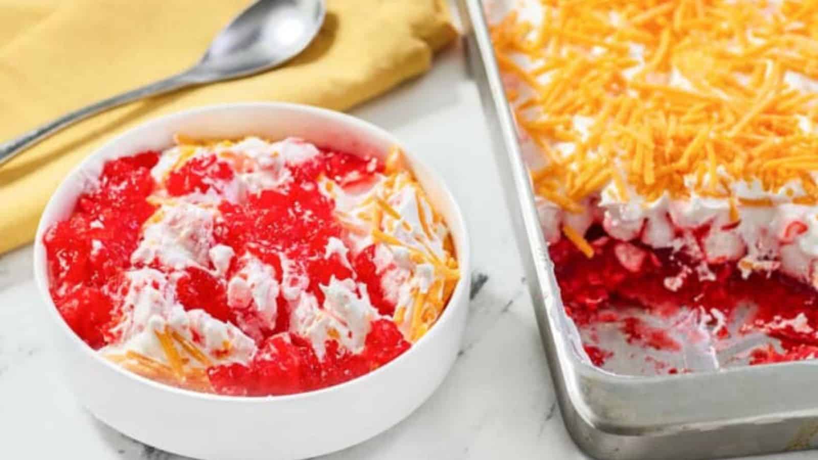 A bowl and pan of red gelatin salad topped with whipped cream and shredded cheddar cheese.