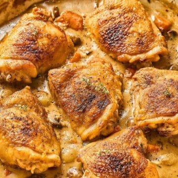 Six golden-brown chicken thighs simmer in a creamy sauce with herbs and vegetables in a skillet.