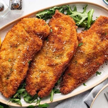 Three breaded chicken cutlets on a platter with greens and lemon wedges, surrounded by tableware and seasonings.