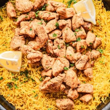 A skillet of yellow rice topped with spiced chicken pieces and lemon wedges, garnished with herbs.