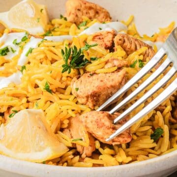 A fork in a bowl of yellow rice with grilled chicken, lemon wedges, herbs, and a dollop of white sauce.