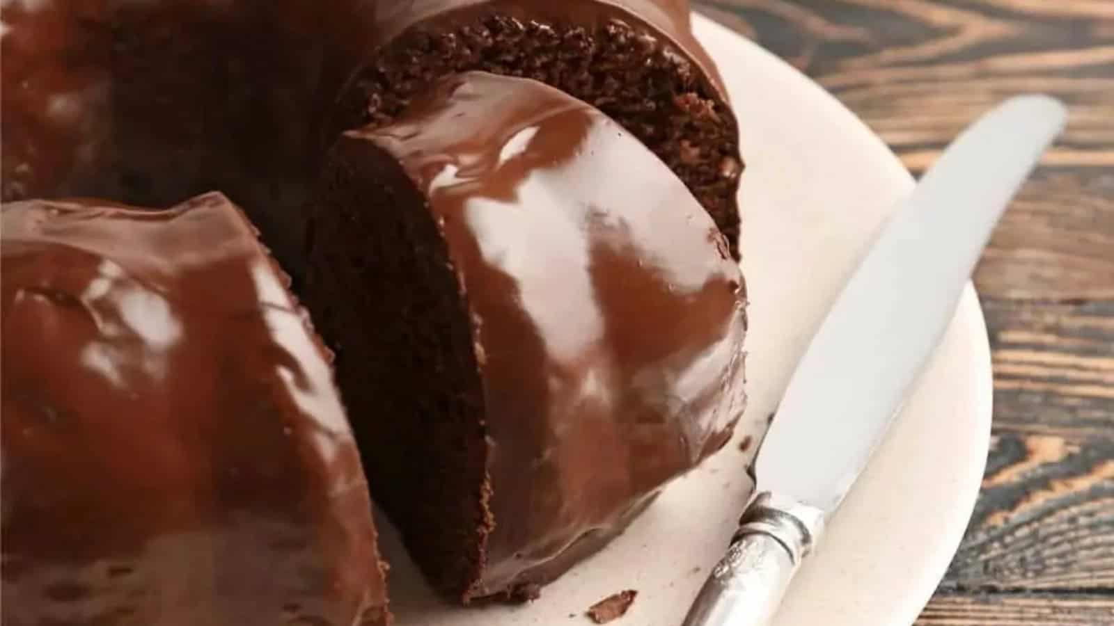 A sliced chocolate bundt cake with glossy chocolate glaze on a plate beside a knife.