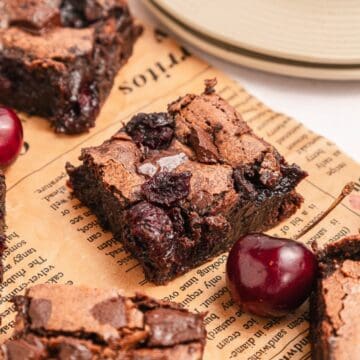 A chocolate cherry brownie on vintage paper, with fresh cherries and other brownie pieces nearby.