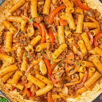 A skillet filled with rigatoni pasta, ground meat, red bell peppers, and sauce, garnished with herbs.