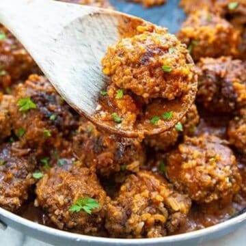 Meatballs in tomato sauce, garnished with herbs, in a skillet; one meatball lifted with a wooden spoon.