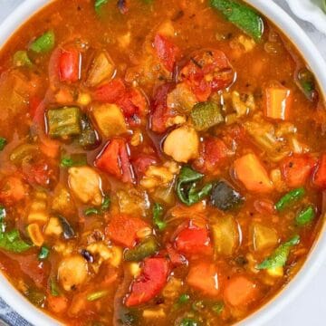A bowl of colorful vegetable soup with tomatoes, carrots, greens, and beans, next to lime wedges and hot sauce.