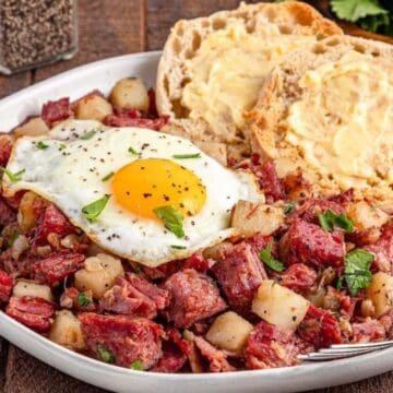 A plate of corned beef hash with diced potatoes, a sunny-side-up egg, and buttered English muffins.