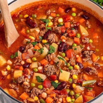 A pot of hearty vegetable and beef soup with beans, peas, carrots, and potatoes, stirred with a wooden spoon.