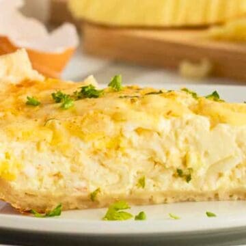 A slice of creamy quiche garnished with parsley on a white plate, with eggs and herbs in the background.