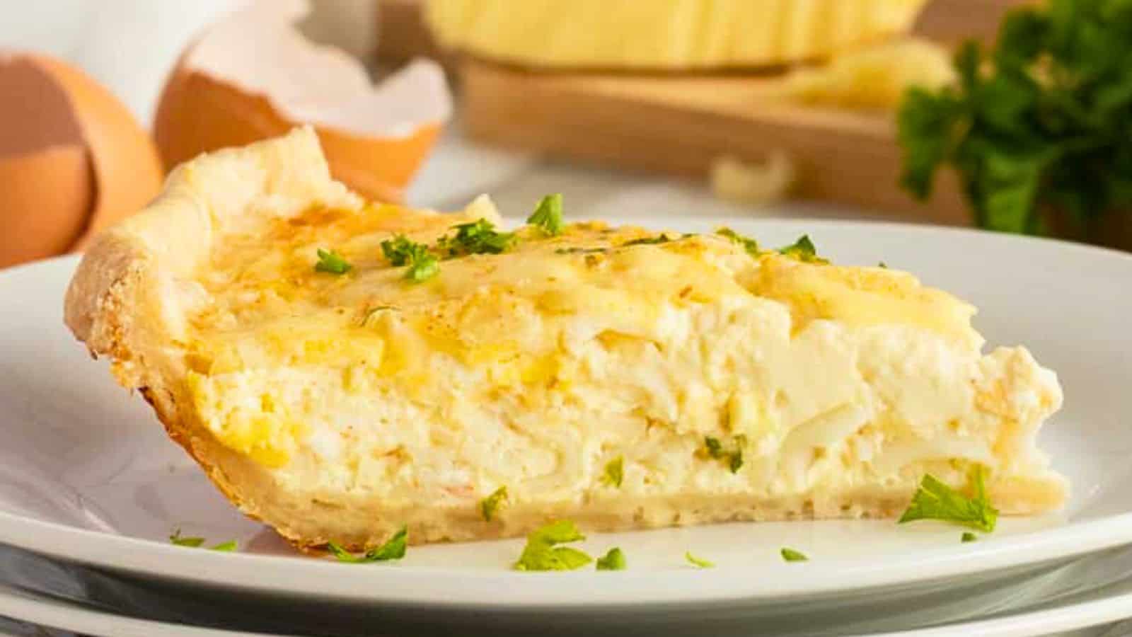 A slice of creamy quiche garnished with parsley on a white plate, with eggs and herbs in the background.