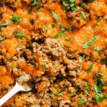 Close-up of cheesy ground beef casserole topped with melted cheese and chopped parsley, with a spoon inside.