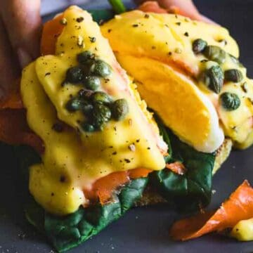 Eggs Benedict with smoked salmon, spinach, capers, and hollandaise sauce on toast, served on a dark plate.