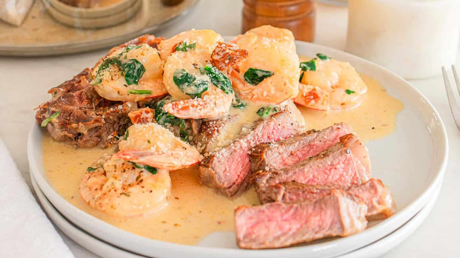 A plate with slices of steak and creamy shrimp in sauce, garnished with spinach.