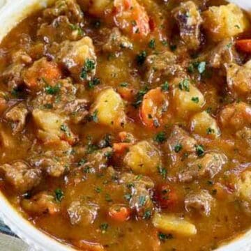 A bowl of beef stew with potatoes, carrots, and herbs, next to a wooden spoon on a striped napkin.