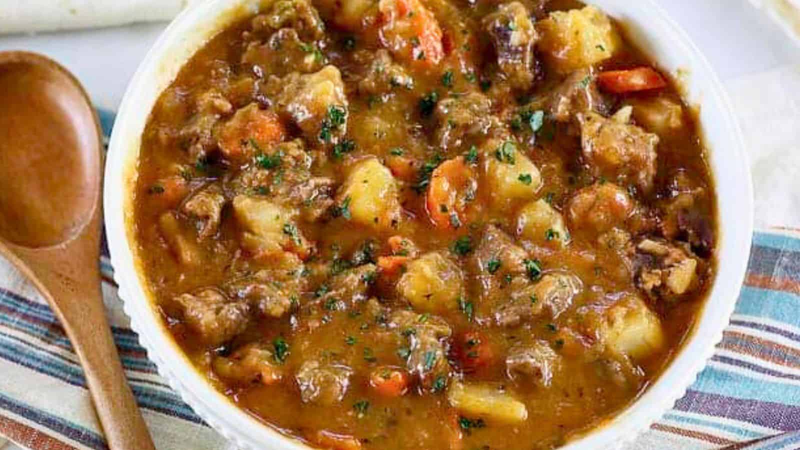 A bowl of beef stew with potatoes, carrots, and herbs, next to a wooden spoon on a striped napkin.