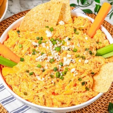 A bowl of buffalo chicken dip garnished with celery sticks, carrot sticks, chips, and sprinkled cheese.