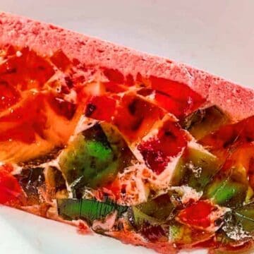 Slice of stained glass jello dessert with colorful gelatin cubes and creamy pink topping on a white plate—a playful nod to forgotten desserts.