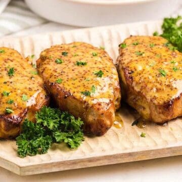 Four grilled pork chops with mustard sauce and parsley on a wooden platter, garnished with fresh parsley.