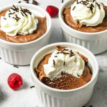 Four ramekins with chocolate mousse topped with whipped cream and chocolate shavings, surrounded by raspberries.