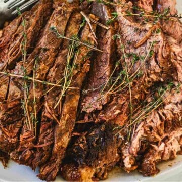 Slices of cooked beef brisket garnished with fresh thyme on a white platter with a serving fork.