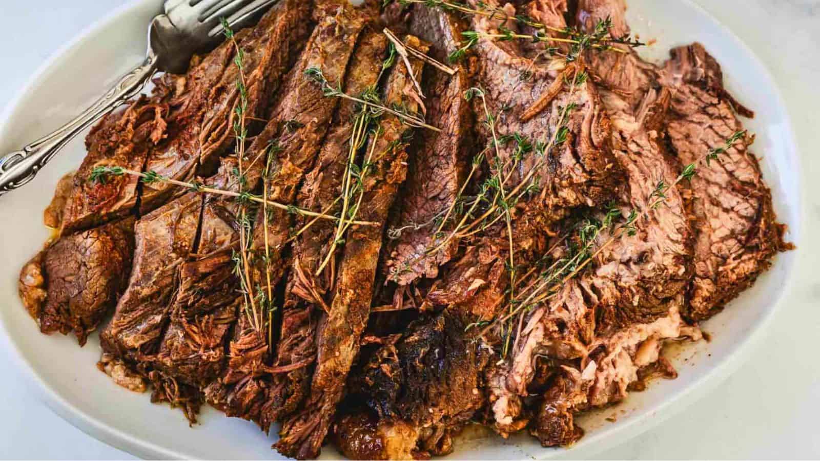 A white oval plate holds several slices of cooked, tender beef roast topped with sprigs of fresh thyme on a light marble surface.