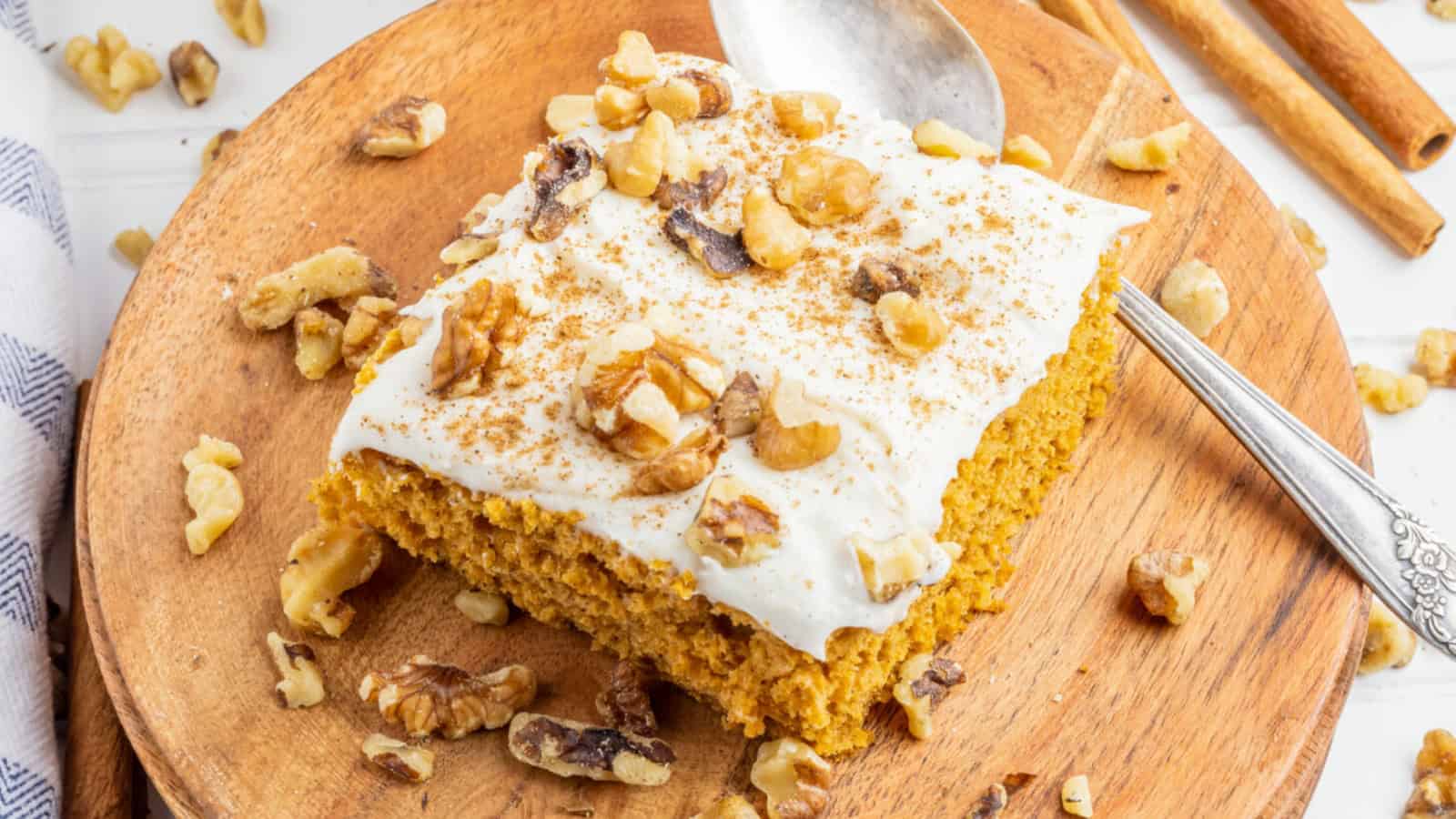 A slice of pumpkin cake with white frosting and walnuts on a wooden plate with a spoon.