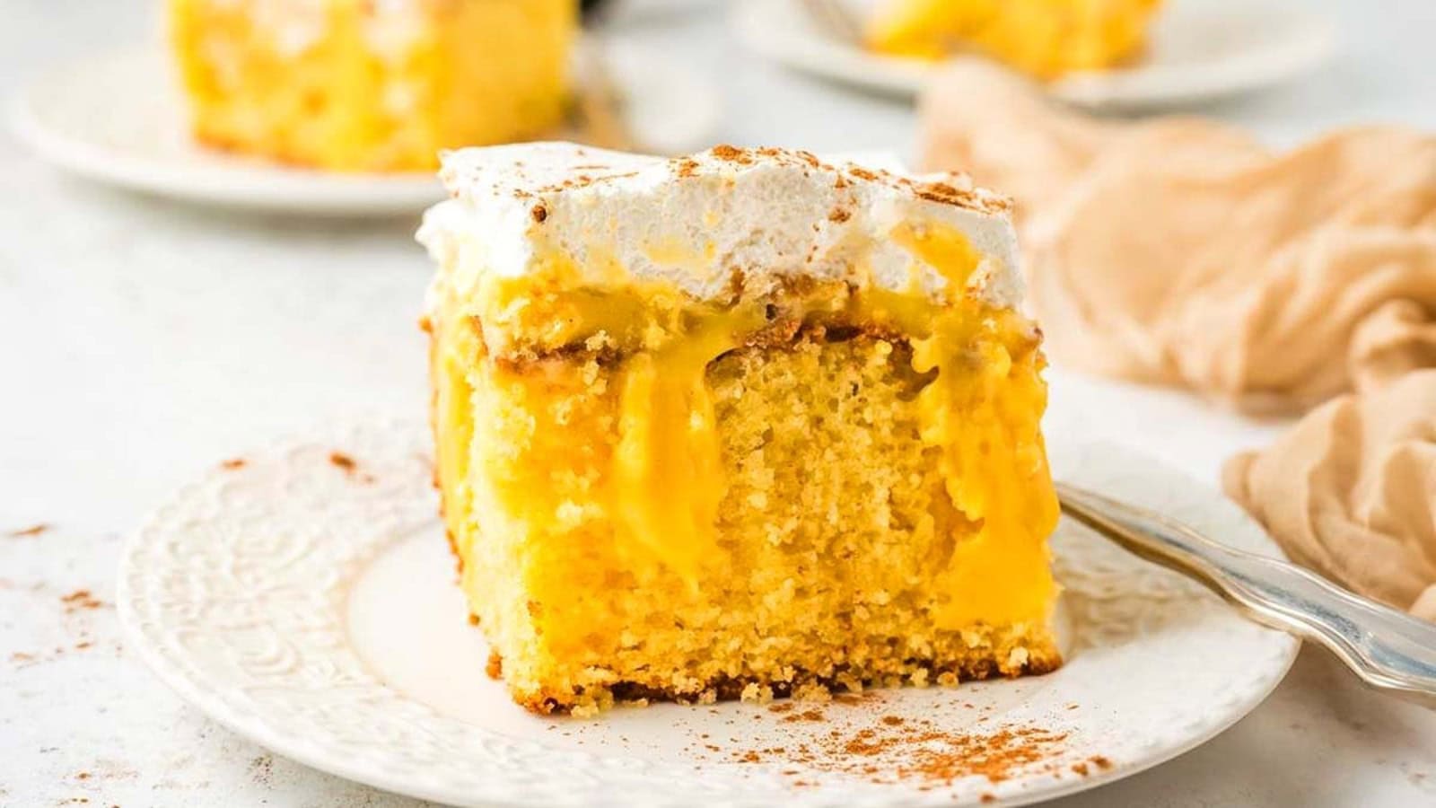 A slice of yellow poke cake with creamy filling and whipped topping on a white plate, with a fork beside it.