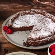 Chocolate cake dusted with powdered sugar, with a slice missing, served on a plate with raspberries.