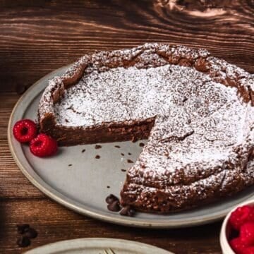 Chocolate cake dusted with powdered sugar, with a slice missing, served on a plate with raspberries.