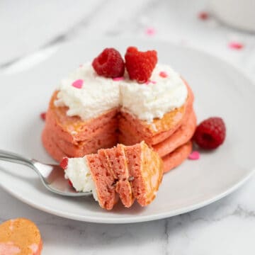 A stack of Raspberry Valentine’s Pancakes with whipped cream and raspberries sits on a white plate; a fork holds a bite.
