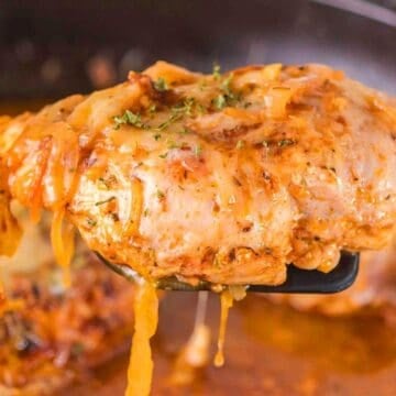 Close-up of cheesy baked chicken with herbs on a spatula, with sauce in a pan in the background.