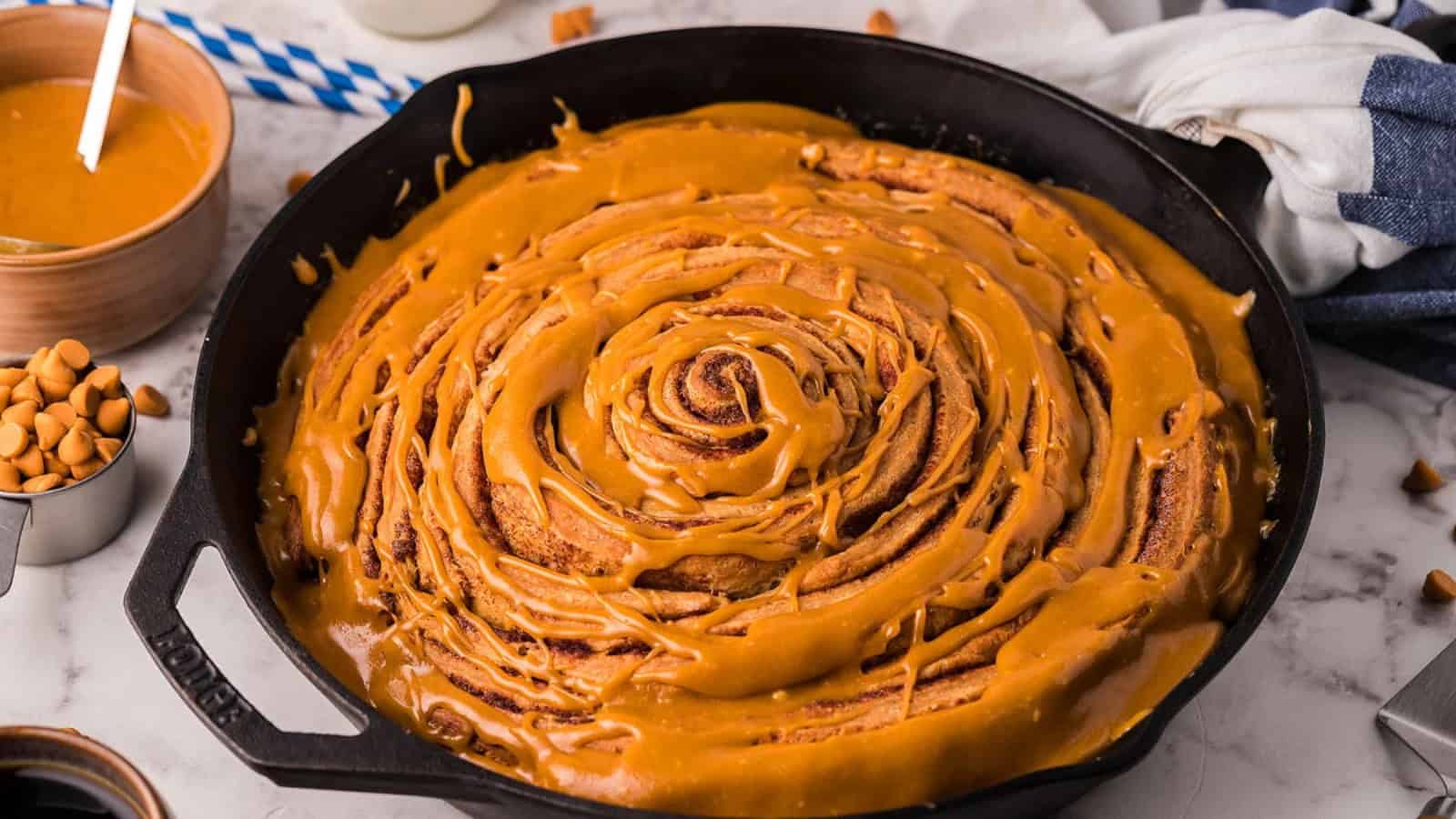A large cinnamon roll with caramel icing in a cast iron skillet, surrounded by baking ingredients.