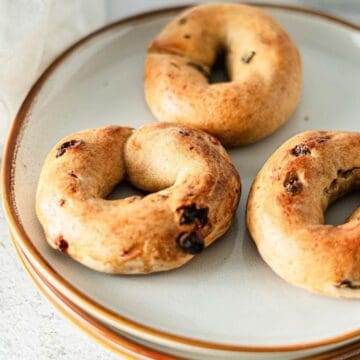 Three homemade bagels with raisins on a white plate, set on a light-colored table.