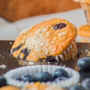Close-up of Cottage Cheese Blueberry Muffins with crumb topping in a baking tin, surrounded by fresh blueberries.