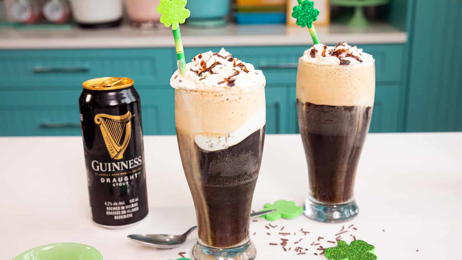 Two Guinness floats topped with whipped cream and chocolate sprinkles, with shamrock straws on a kitchen counter.