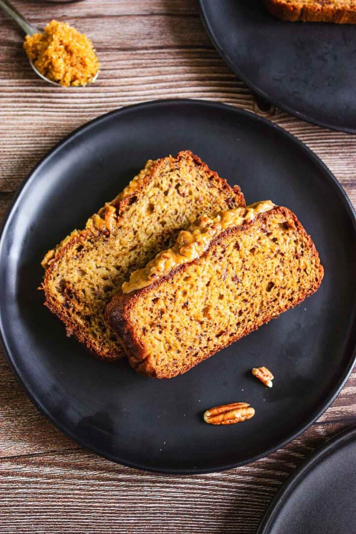 Two slices of banana bread with nuts on a black plate, with a spoon of spread and pecans nearby.