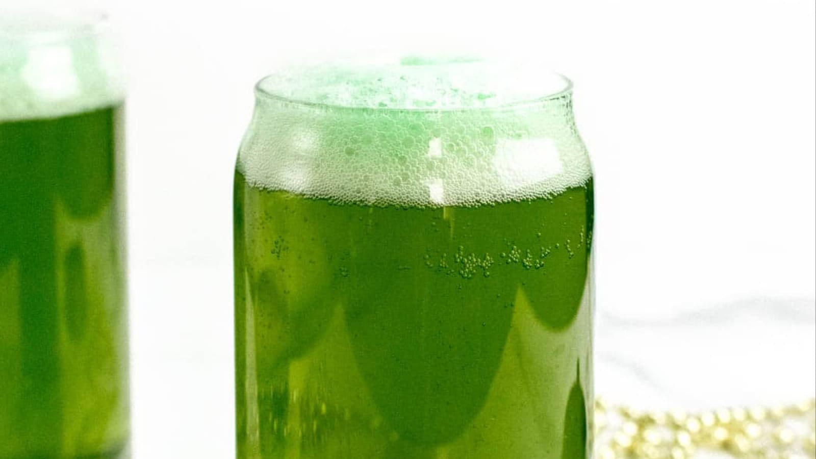 A clear glass of green beer with a foamy top, sitting on a white surface.