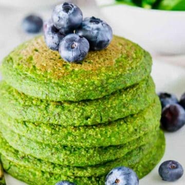 Stack of green pancakes topped with fresh blueberries on a white plate, with a fork beside them.