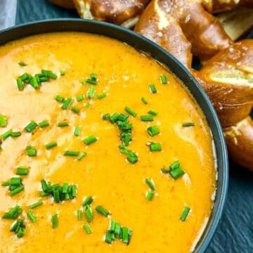 Bowl of beer cheese dip topped with chopped chives, next to soft pretzels on a black plate.