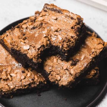 A stack of three chocolate brownies with chocolate chips on a black plate against a light background.