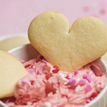 Heart-shaped cookies with pink frosting and sprinkles in a white bowl, surrounded by more cookies.