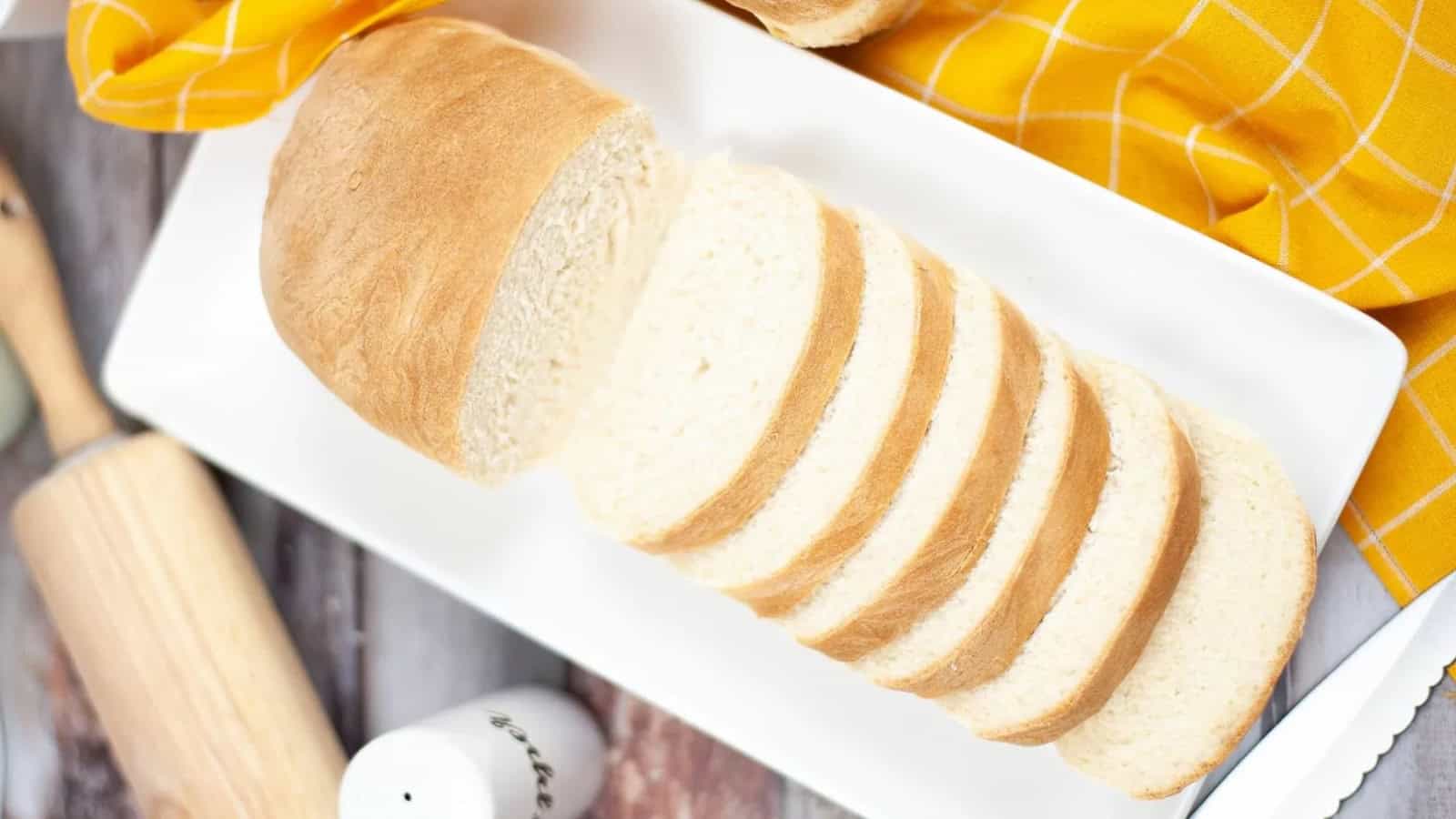 A loaf of sliced white bread on a rectangular white plate, with a yellow plaid cloth in the background.