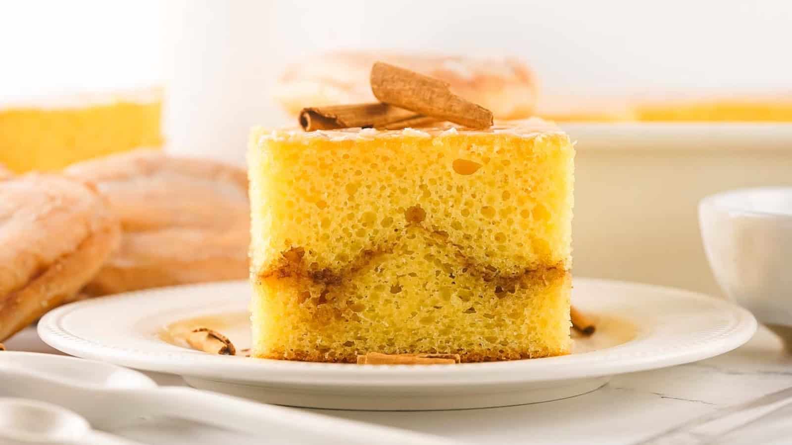 A slice of yellow coffee cake with cinnamon swirl and cinnamon sticks on a white plate.