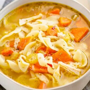 Bowl of chicken noodle soup with carrots, celery, and egg noodles on a striped cloth background.