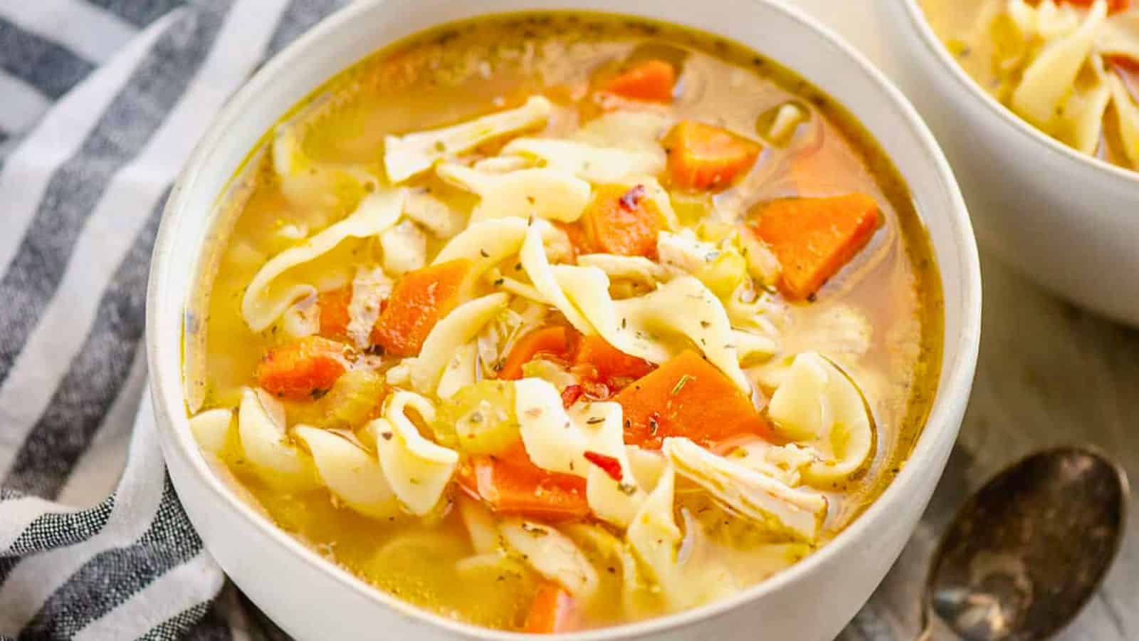 Bowl of chicken noodle soup with carrots, celery, and egg noodles on a striped cloth background.